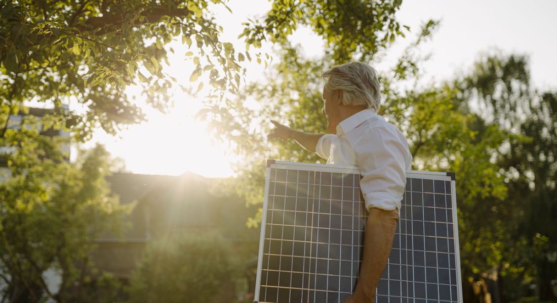 Mann unter einem Baum mit Solarpanel unterm Arm zeigt Richtung Sonne