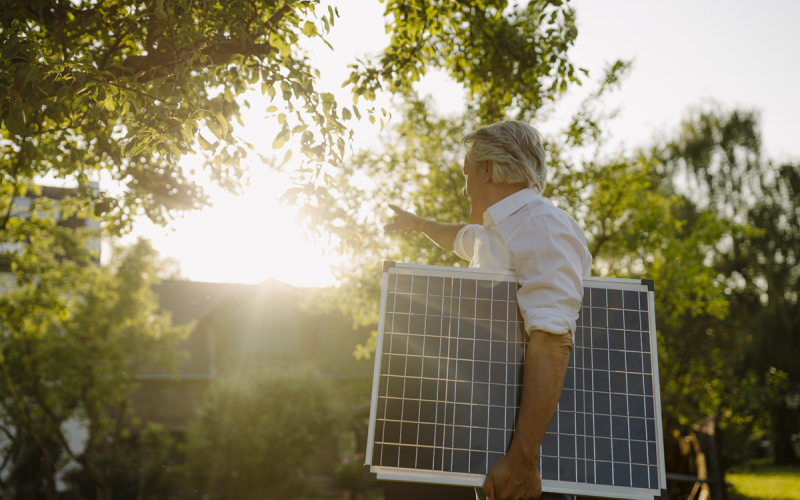 Mann unter einem Baum mit Solarpanel unterm Arm zeigt Richtung Sonne